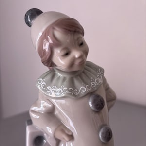 Może przedstawiać: Porcelanowa figurka klauna w beżowym stroju z pasującą czapką, kryzą z białymi wzorami dekoracyjnymi oraz ciemnoszarymi pomponami. Klaun ma brązowe włosy i opiera się o biały blok z szarymi, okrągłymi detalami na powierzchni.