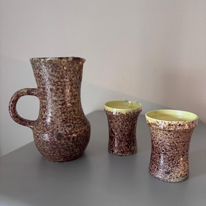 Op de afbeelding: Een bruin gespikkelde keramische kan met een handvat en twee bijpassende bekers. De binnenkant van de bekers is geel. De set heeft een rustieke, handgemaakte uitstraling, geschikt voor het serveren van drankjes.