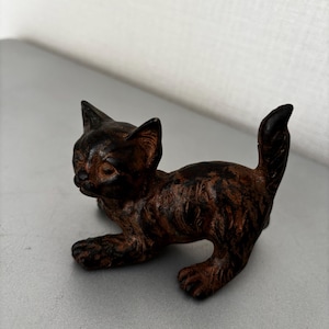 May include: A small, dark brown, textured metal figurine of a kitten in a playful, crouching stance with its tail held upright. The surface of the cat sculpture features a weathered, mottled finish with darker and lighter brown tones throughout.