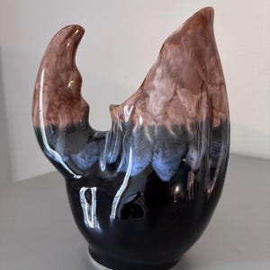 May include: A ceramic vase with an unusual, organic form. The vase displays a colour gradient, shifting from a dark black base to blues, browns, and a lighter brown at the top. The design incorporates a curved, handle-like feature.