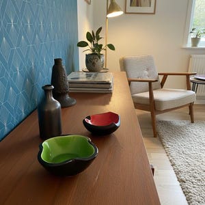 May include: An interior shot showcasing a wooden table with various decorative objects. Two small, black bowls with red and green interiors are on the table. A dark bottle and a textured vase are also present. A lamp and armchair are in the background.