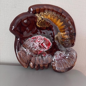 May include: A unique, ceramic decorative dish with a glossy, brown glaze. The dish features a textured design with accents of red, yellow, and white. The shape is organic and asymmetrical, adding to its artistic appeal.