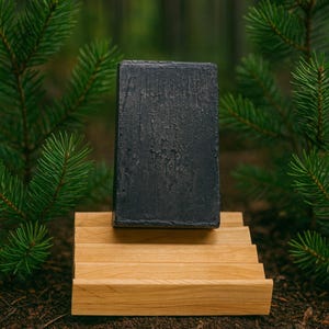 Può includere: Una saponetta rettangolare grigio scuro poggia su un portasapone in legno marrone chiaro. Il sapone ha una superficie strutturata. Rami di pino verdi incorniciano il sapone e il portasapone, su uno sfondo sfocato.