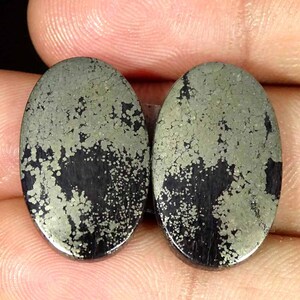 May include: Two oval-shaped cabochons featuring a metallic, brassy gold pyrite inclusion pattern set against a dark, opaque black background. The stones have a smooth, polished surface with a reflective finish and distinct, irregular mineral speckling.