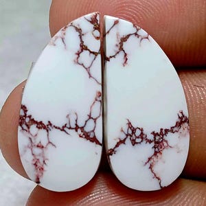 May include: Two teardrop-shaped white gemstones featuring a distinct network of dark reddish-brown veining throughout the surface. The stones are polished to a smooth finish and exhibit a high-contrast pattern of white and deep brown lines.