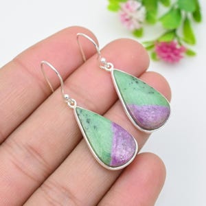 May include: A pair of teardrop-shaped drop earrings featuring polished zoisite stones with distinct green and purple colour zones. Each stone is set in a simple silver-toned metal bezel with a matching silver-toned hook for ear attachment.