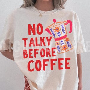 Può includere: T-shirt color crema con la scritta rossa "NO TALKY BEFORE COFFEE". A destra c'è una grafica colorata di una caffettiera. La caffettiera è arancione, rosa e blu con dettagli floreali. È visibile una collana d'oro.