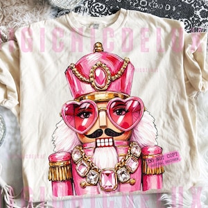 Può includere: Una t-shirt color crema con un'illustrazione grafica di uno schiaccianoci che indossa un'uniforme rosa, una corona abbinata con una gemma centrale e occhiali rosa a forma di cuore. Lo schiaccianoci ha i baffi neri e una grande collana gioiello.