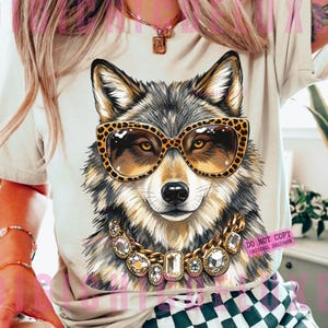 May include: A beige t-shirt featuring a graphic illustration of a wolf wearing leopard print sunglasses and a gold chain necklace with large clear crystals. The wolf has grey, black, and white fur, and the design is centered on the front of the shirt.
