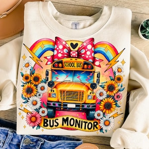 May include: Cream-colored sweatshirt featuring a vibrant school bus graphic. The design includes a yellow school bus, a large polka-dot bow, sunflowers, daisies, rainbows, and the text "SCHOOL BUS" and "BUS MONITOR".