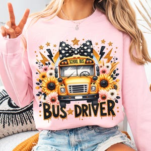 May include: Pink sweatshirt with a yellow school bus graphic, a black and white polka dot bow, and the text "BUS DRIVER" in black with gold glitter accents.
