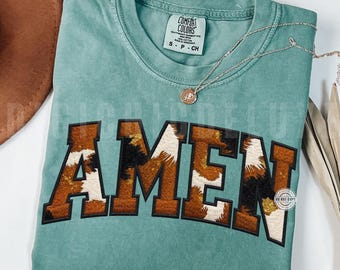 Könnte beinhalten: Ein türkisgrünes T-Shirt mit dem Wort "AMEN" in einem braunen und weißen Kuhdruck-Design. Das Shirt hat einen Rundhalsausschnitt und kurze Ärmel. Eine silberne Halskette mit Anhänger ist über dem Shirt drapiert.