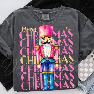 Hand-painted watercolor nutcracker soldier Christmas PNG design in vibrant pink, gold, and blue with festive stars and layered “Merry Christmas” lettering. Perfect for holiday shirts, Christmas party outfits, Nutcracker ballet gifts, winter home décor, festive wall art, seasonal apparel, cozy holiday decorations, and Christmas gift ideas for women and kids.