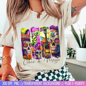 May include: A white t-shirt with a colorful graphic design featuring a guitar, a margarita glass, and sugar skulls. The text "Cinco de Mayo" is printed on the shirt.