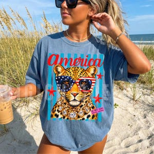 Patriotic leopard PNG design featuring glamorous leopard wearing American flag sunglasses and chunky gold jewelry on retro blue striped background. Trendy USA coquette animal graphic perfect for 4th of July shirts, patriotic apparel, summer vacation tees, western glam fashion, Independence Day party outfit, leopard lover gift, sublimation PNG, DTF transfer, print on demand design, tumbler wrap, mug design, wall art printable, boutique graphic tee, trendy Americana aesthetic clipart.