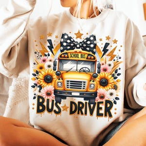 May include: A white sweatshirt with a yellow school bus graphic, a black and white polka dot bow, sunflowers, and the text "BUS DRIVER" in gold glitter.