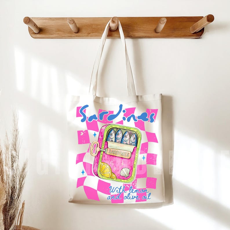 Trendy sardines shirt design featuring a pink illustrated sardine tin can with fish, lemon slices, shell detail, blue lettering, and hot pink checkerboard background. Cute Mediterranean foodie graphic perfect for coastal Europe vacations, European summer outfits design, fish market aesthetic, seafood lover gifts, retro vacation shirts design, beach trip apparel design, coastal boutique fashion, and trendy graphic shirt design for women, girls trips, and summer travel. For sublimation and DTF.