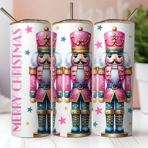 May include: A set of three white stainless steel tumblers featuring a pink and blue nutcracker design. The nutcracker wears a pink hat and uniform with gold accents. The words "MERRY CHRISTMAS" are printed vertically in pink. Blue and gold stars are scattered around the design.