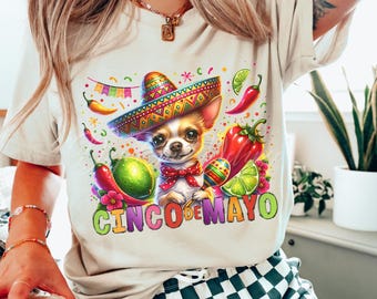 Puede incluir: Una camiseta blanca con una ilustración colorida de un chihuahua con un sombrero y sosteniendo maracas. El texto "Cinco de Mayo" está escrito en una fuente colorida debajo del chihuahua.