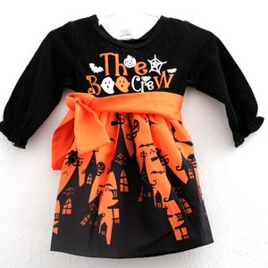 Op de afbeelding: Een zwarte jurk met lange mouwen en de tekst The Boo Crew met witte en oranje Halloween-iconen. De rok heeft een oranje tailleband en een zwart-oranje print van spookhuizen, vleermuizen en heksen. De mouwen hebben geplooide boorden.