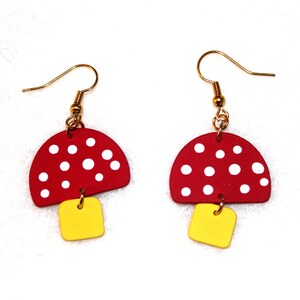May include: A pair of red and white mushroom-shaped earrings with yellow accents. The earrings are made of wood and have a gold-tone finish.