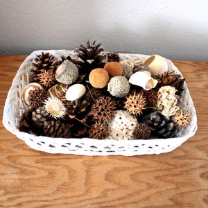 May include: A white wicker basket filled with a variety of natural items, including pinecones, acorns, and dried flowers. The basket is sitting on a wooden surface.