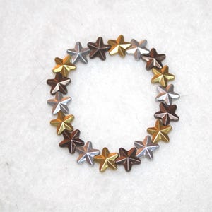 Puede incluir: Un círculo de cuentas con forma de estrella en tonos plateado, dorado y marrón.