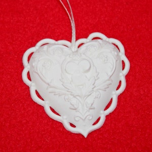May include: White ceramic heart ornament with a detailed floral design. The heart has a scalloped edge and a loop for hanging.