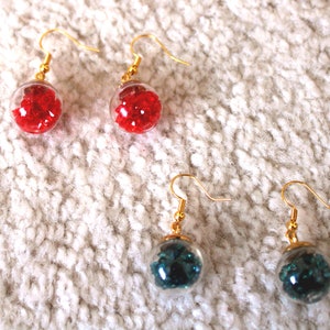 Puede incluir: Un par de pendientes de aro dorados con esferas de vidrio transparentes llenas de purpurina roja y azul verdosa.