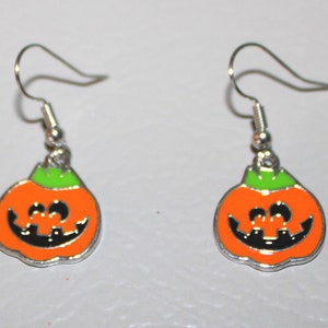 Könnte beinhalten: Ein Paar silberne Ohrhänger mit orangefarbenen und grünen Kürbisanhängern. Die Kürbisse haben schwarze Augen, eine Nase und einen lächelnden Mund.