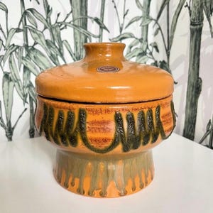 Op de afbeelding: Een vintage keramische pot met deksel met een bruine, groene en oranje glazuur. De pot heeft een uniek ontwerp met een golvend patroon rond het midden en een druppelglazuureffect op de basis.