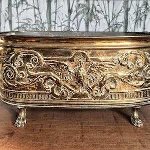 Könnte beinhalten: Ein rechteckiger, antiker Messing-Pflanzkübel mit aufwendigen Reliefdetails. Der Pflanzkübel weist ein dekoratives Design mit Vögeln und Wirbelmustern auf, das von Klauenfüßen getragen wird. Er hat Ringgriffe und ein dunkles Inneres.