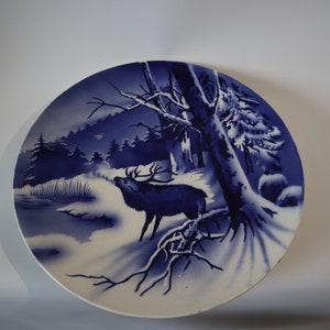 Könnte beinhalten: Ein blauer und weißer Keramikteller mit einer Winterszene, die ein Reh in einem schneebedeckten Wald zeigt. Das Reh steht im Vordergrund, im Hintergrund sind Bäume und schneebedeckter Boden zu sehen.