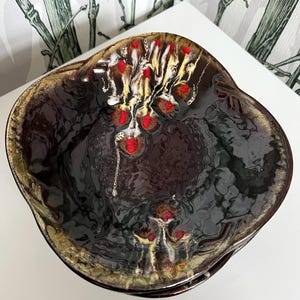 Op de afbeelding: Een bruine keramische kom met een gegolfde rand en een abstract ontwerp in zwart, wit en rood. De kom heeft een glanzende afwerking.