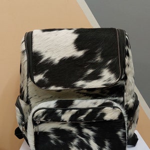 May include: A black and white cowhide backpack with a zippered front pocket. The backpack has a black leather trim and a black zipper.