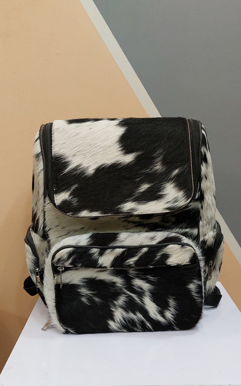 May include: A black and white cowhide backpack with a zippered front pocket. The backpack has a black leather trim and a black zipper.
