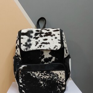 May include: A black and white cowhide backpack with a black leather trim and zipper accents. The backpack has a large main compartment and two smaller pockets on the front.