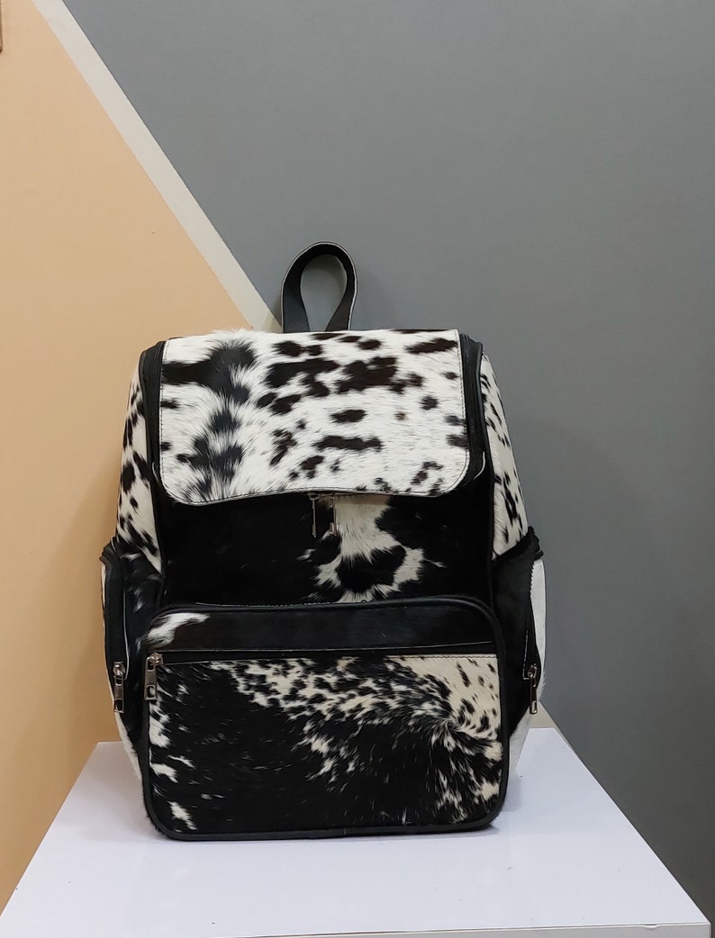 May include: A black and white cowhide backpack with a black leather trim and zipper accents. The backpack has a large main compartment and two smaller pockets on the front.