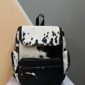 May include: Black and white cowhide backpack with a large front pocket and zipper closures.