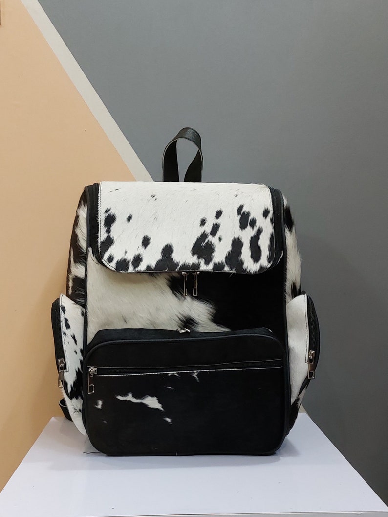 May include: Black and white cowhide backpack with a large front pocket and zipper closures.