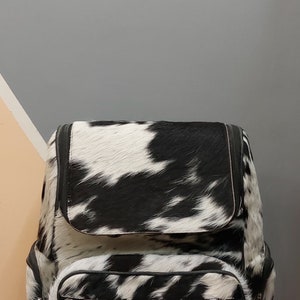 May include: Black and white cowhide backpack with a large main compartment and a smaller front pocket. The backpack has a zipper closure and adjustable straps.