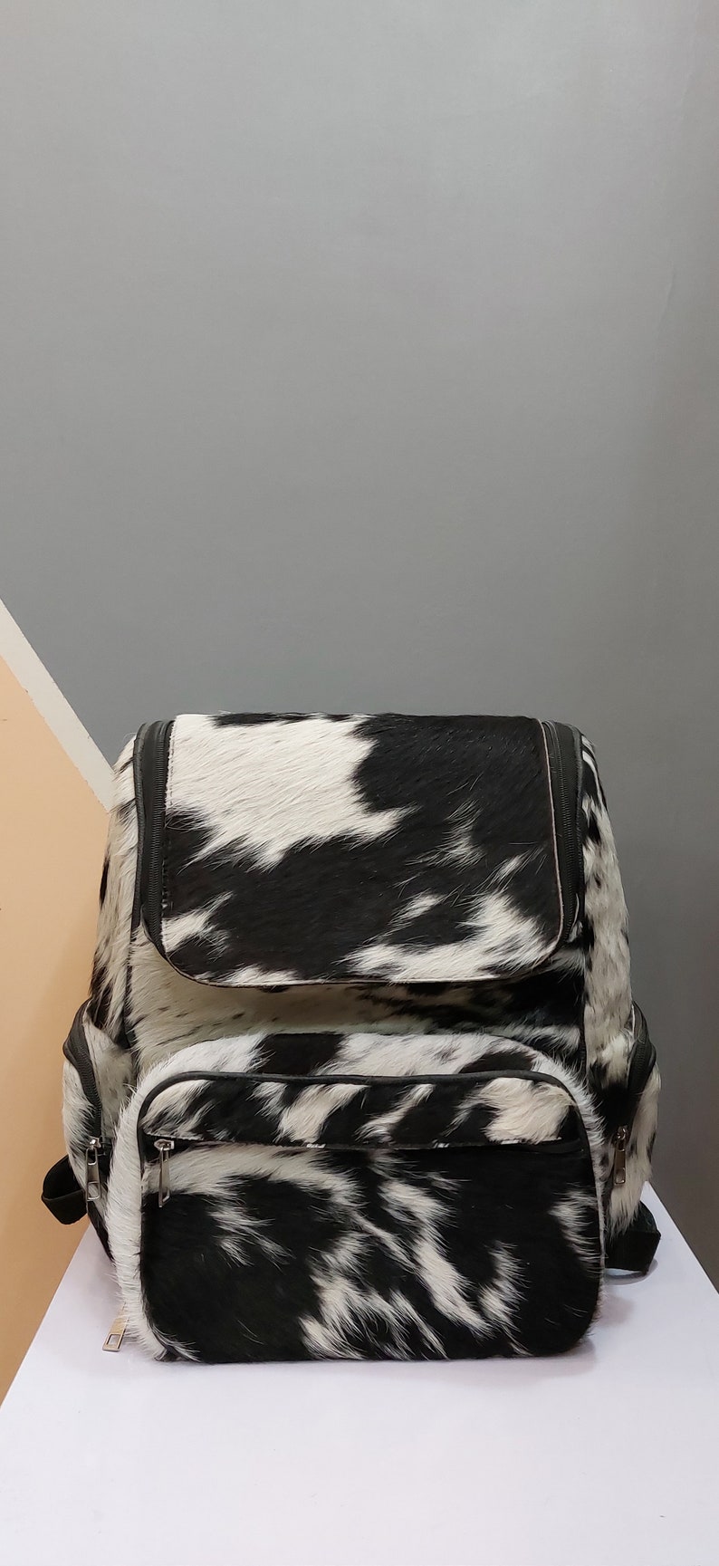 May include: Black and white cowhide backpack with a large main compartment and a smaller front pocket. The backpack has a zipper closure and adjustable straps.