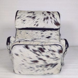 May include: A white and black cowhide leather backpack with two zippered compartments and a top flap closure.