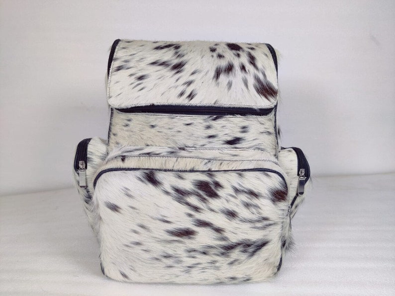 May include: A white and black cowhide leather backpack with two zippered compartments and a top flap closure.