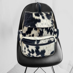 May include: A black and white cowhide backpack with a front zippered pocket. The backpack is sitting on a black chair.