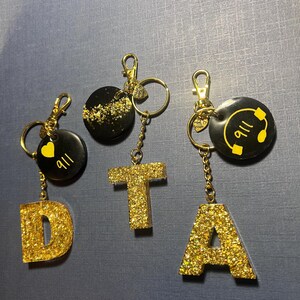 May include: Four gold glitter letter keychains, spelling out "DTA", with two additional black and gold keychains. One keychain has a heart with "all" written on it, and the other has a phone icon with "911" written on it.