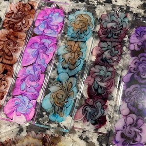 Könnte beinhalten: Fünf Acryl-Lesezeichen mit floralem Design. Die Lesezeichen sind transparent und zeigen ein wirbelndes Muster aus brauner, rosa, blauer, violetter und türkisfarbener Farbe.