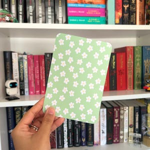 Può includere: Una carta rettangolare con angoli arrotondati, con un motivo di piccoli fiori bianchi su sfondo verde chiaro. La carta è tenuta davanti a una libreria piena di libri.