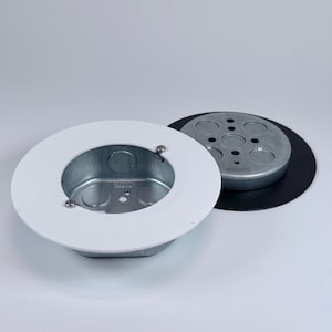 May include: A metal electrical box with a white trim ring and a separate metal cover. The box has a square shape with rounded corners and is designed for recessed installation. The cover is round and has multiple holes.