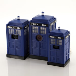 May include: Three blue Police Box models of different sizes. Each box has a black base, a sign reading "POLICE BOX", and a light on top. The boxes feature windows and a door with a circular detail.